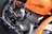 A close-up view of a motorcycle engine, prominently displaying a polished metallic finish. The frame and elements around the engine are colored in bright orange. The motorcycle brand name is visible on the orange part of the frame. The engine's design includes visible coils and exhaust pipes.