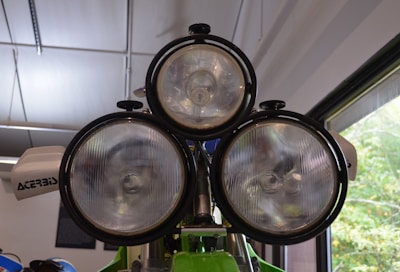 Three large, circular headlights are mounted on a vehicle. Two headlights are side-by-side below a third one centered above them. The headlights have clear, ribbed glass covers, and the vehicle appears to be indoors, possibly in a showroom or workshop. The surrounding area has white walls, a ceiling with parallel light fixtures, and a large window on the right allowing natural light in. A white accessory with the word 'ACERBIS' is visible on the left.