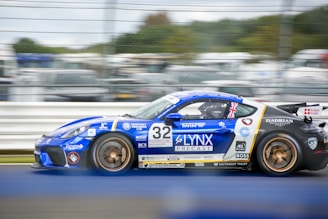 A blue sports car with detailed sponsor logos and markings is racing on a track. The car features a Union Jack flag near its rear window, and the number 32 is prominently displayed on its side. The background shows blurred spectators and other cars, indicating motion and speed.