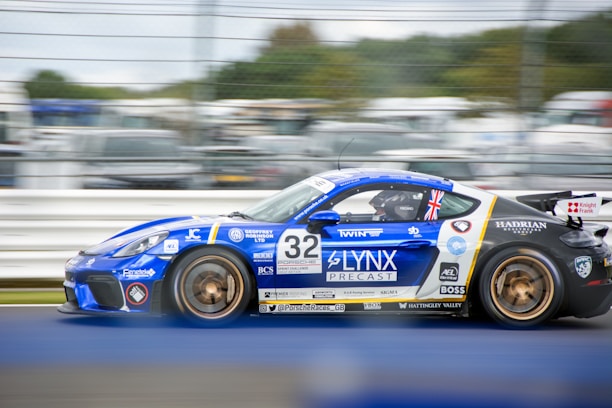 A blue sports car with detailed sponsor logos and markings is racing on a track. The car features a Union Jack flag near its rear window, and the number 32 is prominently displayed on its side. The background shows blurred spectators and other cars, indicating motion and speed.
