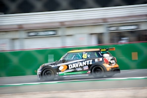 A racing car is speeding on a track, featuring a black body with colorful decals, including logos and the number 45. The background is blurred, indicating fast motion. Sponsorships from various brands are visible on the car's body.