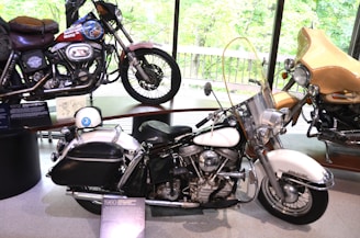 A display of three vintage motorcycles inside a museum with large windows providing a view of greenery outside. The motorcycles are of different styles and colors, featuring intricate detailing and chrome finishes. Each bike is accompanied by informational plaques highlighting their models and history.