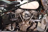 Close-up view of a vintage motorcycle engine with metallic components, including a shiny chrome air filter cover reflecting its surroundings. The engine displays intricate mechanical details with visible cylinders, pipes, and other parts. The seat and part of the fuel tank are also visible.