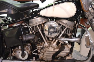 Close-up view of a vintage motorcycle engine with metallic components, including a shiny chrome air filter cover reflecting its surroundings. The engine displays intricate mechanical details with visible cylinders, pipes, and other parts. The seat and part of the fuel tank are also visible.