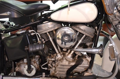 Close-up view of a vintage motorcycle engine with metallic components, including a shiny chrome air filter cover reflecting its surroundings. The engine displays intricate mechanical details with visible cylinders, pipes, and other parts. The seat and part of the fuel tank are also visible.