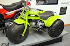 A bright green three-wheeled all-terrain vehicle with large, thick tires is prominently displayed next to a red and black CB125T motorcycle. The green vehicle features a compact engine and is stationed on a platform, surrounded by informational panels discussing motorcycle history.
