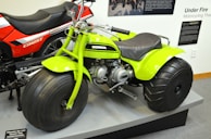 A bright green three-wheeled all-terrain vehicle with large, thick tires is prominently displayed next to a red and black CB125T motorcycle. The green vehicle features a compact engine and is stationed on a platform, surrounded by informational panels discussing motorcycle history.
