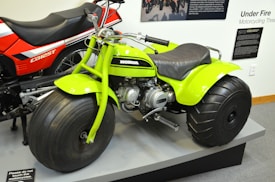 A bright green three-wheeled all-terrain vehicle with large, thick tires is prominently displayed next to a red and black CB125T motorcycle. The green vehicle features a compact engine and is stationed on a platform, surrounded by informational panels discussing motorcycle history.