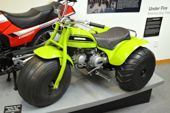 A bright green three-wheeled all-terrain vehicle with large, thick tires is prominently displayed next to a red and black CB125T motorcycle. The green vehicle features a compact engine and is stationed on a platform, surrounded by informational panels discussing motorcycle history.