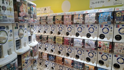 A brightly lit wall filled with rows of capsule toy vending machines, each featuring various toy or character themes. The machines have transparent windows displaying images of possible collectible items. Some machines have pricing labels in yen and accompanying signage with Japanese text.