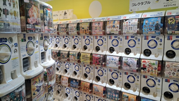 A brightly lit wall filled with rows of capsule toy vending machines, each featuring various toy or character themes. The machines have transparent windows displaying images of possible collectible items. Some machines have pricing labels in yen and accompanying signage with Japanese text.