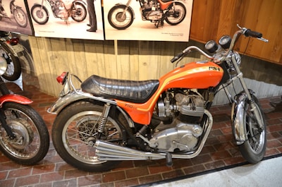 A classic orange motorcycle with 750 CC engine displacement is displayed indoors, surrounded by other vintage motorcycles. It has a black leather seat, chrome accents, and two round gauges on the handlebars. The setting appears to be a museum or exhibit with wooden and concrete walls.