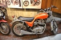 A classic orange motorcycle with 750 CC engine displacement is displayed indoors, surrounded by other vintage motorcycles. It has a black leather seat, chrome accents, and two round gauges on the handlebars. The setting appears to be a museum or exhibit with wooden and concrete walls.