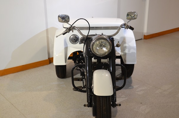 A vintage three-wheeled motorcycle with a large headlight and chrome handlebars. The motorcycle has white fenders and a white back, with black elements in the body and frame.