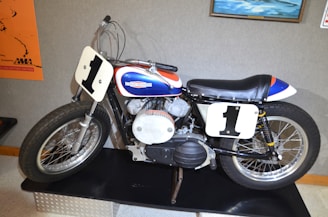 A vintage motorcycle with a white number plate displaying the number '1' on both front and side. The motorcycle has a sleek black seat and a vibrant color scheme featuring blue, red, and white. It is displayed on a platform, likely in a museum or exhibition setting, with posters and paintings visible in the background.