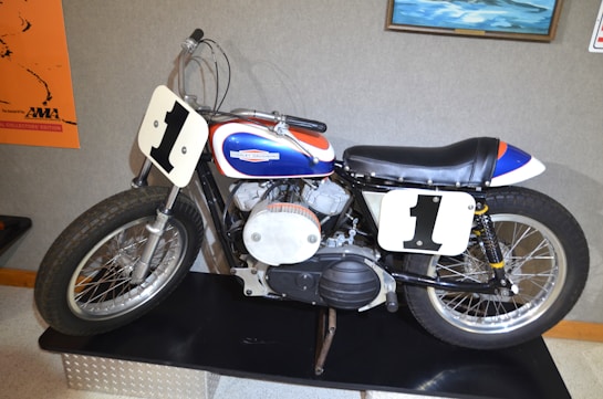 A vintage motorcycle with a white number plate displaying the number '1' on both front and side. The motorcycle has a sleek black seat and a vibrant color scheme featuring blue, red, and white. It is displayed on a platform, likely in a museum or exhibition setting, with posters and paintings visible in the background.