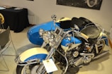 A vintage motorcycle with a sidecar is displayed indoors, featuring a classic design with a prominent front fender and headlights. The motorcycle is painted in blue and cream, with chrome details on the engine and handlebars. Nearby, there is a black-covered table with various items, likely papers or brochures.