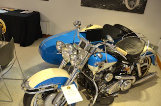 A vintage motorcycle with a sidecar is displayed indoors, featuring a classic design with a prominent front fender and headlights. The motorcycle is painted in blue and cream, with chrome details on the engine and handlebars. Nearby, there is a black-covered table with various items, likely papers or brochures.