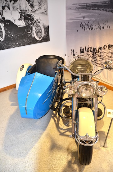 A vintage motorcycle with a sidecar is displayed indoors on a polished floor. The sidecar is painted in a bright blue color, while the motorcycle itself is primarily white with chrome accents. In the background, there are vintage black and white photographs of people and another motorcycle, adding to the historical ambiance.