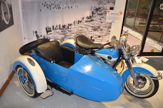 A vintage motorcycle with a sidecar is parked indoors on a tiled floor. The motorcycle and sidecar are painted blue and white, with chrome details and black leather seats. A large black and white photograph in the background depicts a crowd of people and a line of vintage cars. There is a glass door to the right, leading outside.