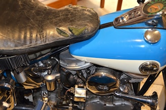 A close-up view of a Harley-Davidson motorcycle showing a worn-out black leather seat, a blue and white fuel tank with a circular emblem, and the shiny chrome parts of the engine.
