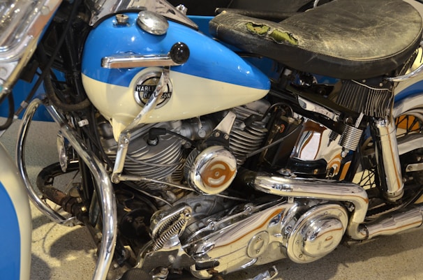 A close-up view of a vintage motorcycle featuring a Harley-Davidson logo on the blue and cream gas tank. The motorcycle has a worn black leather seat and polished chrome engine parts.