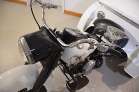 The image features a close-up of a classic motorcycle with a black and white color scheme. The front view highlights the handlebars, headlight, and part of the engine. A sidecar is attached to the motorcycle, with a black seat and a chrome rearview mirror visible.