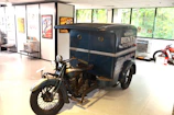 A vintage motorcycle with a sidecar is displayed in what appears to be a museum or showroom. The sidecar features signage indicating a cycle shop and delivery service. The surroundings include framed posters and memorabilia related to motorcycles, suggesting an exhibition space.