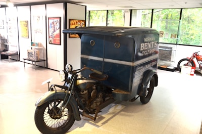 A vintage motorcycle with a sidecar is displayed in what appears to be a museum or showroom. The sidecar features signage indicating a cycle shop and delivery service. The surroundings include framed posters and memorabilia related to motorcycles, suggesting an exhibition space.