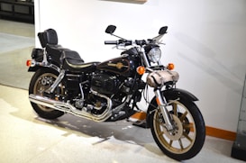 A classic-style black motorcycle with chrome elements and a leather padded seat is parked indoors. It features a vintage design with a round headlight, leather saddlebags, and an emblem on the fuel tank. The motorcycle is displayed on a light-colored floor against a white wall, with its side mirrors and handlebars clearly visible.