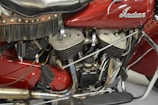 A close-up view of a vintage motorcycle engine with intricate mechanical details and a leather seat with fringe. The surface of the engine parts is metallic and slightly worn, suggesting age and use.
