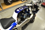 A motorcycle with the fairing removed, revealing the internal components such as the engine and parts of the fuel system. The bike is mostly blue and white with a black seat, parked inside a garage or workshop space with a cement floor. A shelf and another motorcycle are visible in the background.