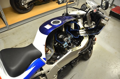 A motorcycle with the fairing removed, revealing the internal components such as the engine and parts of the fuel system. The bike is mostly blue and white with a black seat, parked inside a garage or workshop space with a cement floor. A shelf and another motorcycle are visible in the background.