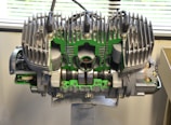A large, detailed mechanical engine model is displayed indoors. The engine's components are exposed and highlighted with green accents. Metallic fins are prominent at the top of the structure, indicating cooling mechanisms. The background features a partially visible window with blinds, suggesting a laboratory or workshop setting.