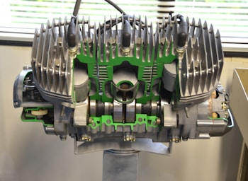 A large, detailed mechanical engine model is displayed indoors. The engine's components are exposed and highlighted with green accents. Metallic fins are prominent at the top of the structure, indicating cooling mechanisms. The background features a partially visible window with blinds, suggesting a laboratory or workshop setting.