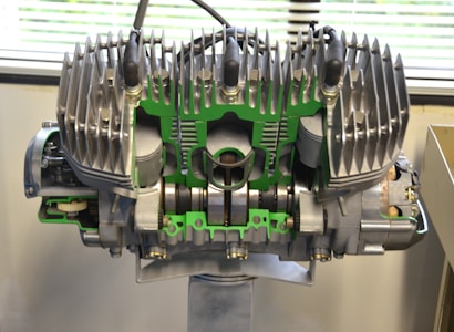 A large, detailed mechanical engine model is displayed indoors. The engine's components are exposed and highlighted with green accents. Metallic fins are prominent at the top of the structure, indicating cooling mechanisms. The background features a partially visible window with blinds, suggesting a laboratory or workshop setting.