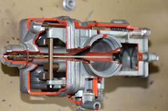 A detailed cross-sectional view of a mechanical component, likely a carburetor, is displayed. The interior structures and mechanisms are visible, showcasing various chambers, passages, and metal parts. The metal surfaces are complemented by orange-colored highlights to emphasize specific sections.