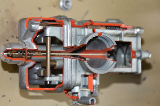 A detailed cross-sectional view of a mechanical component, likely a carburetor, is displayed. The interior structures and mechanisms are visible, showcasing various chambers, passages, and metal parts. The metal surfaces are complemented by orange-colored highlights to emphasize specific sections.