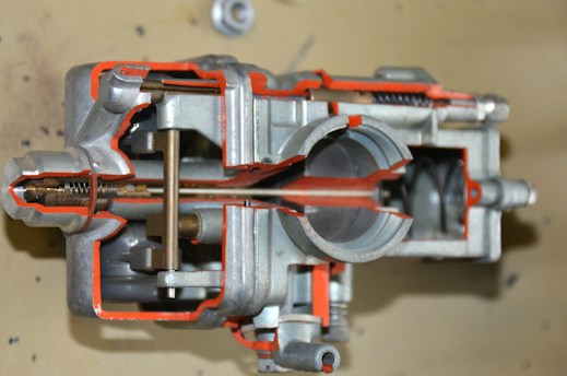 A detailed cross-sectional view of a mechanical component, likely a carburetor, is displayed. The interior structures and mechanisms are visible, showcasing various chambers, passages, and metal parts. The metal surfaces are complemented by orange-colored highlights to emphasize specific sections.