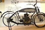 An antique motorcycle with a black metal frame and visible engine components sits on a stand. Behind it, detailed technical drawings are displayed, showcasing engine parts and components.