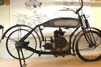 An antique motorcycle with a black metal frame and visible engine components sits on a stand. Behind it, detailed technical drawings are displayed, showcasing engine parts and components.