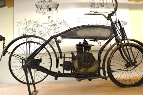 An antique motorcycle with a black metal frame and visible engine components sits on a stand. Behind it, detailed technical drawings are displayed, showcasing engine parts and components.