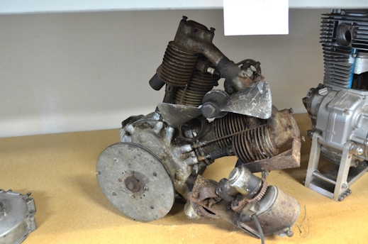 A collection of aged, rusty engine parts is placed on a wooden surface, including a metal disk and cylindrical components with visible wear and tear. The engine components exhibit signs of oxidation and corrosion, showcasing a historical or vintage appearance.