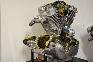 A sectioned motorcycle engine is displayed with detailed internal components visible. The machinery includes gears, pistons, and other mechanical parts, highlighted with bright yellow and blue accents to emphasize certain areas. The engine is mounted on a stand against a plain background.