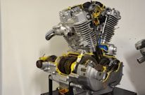 A sectioned motorcycle engine is displayed with detailed internal components visible. The machinery includes gears, pistons, and other mechanical parts, highlighted with bright yellow and blue accents to emphasize certain areas. The engine is mounted on a stand against a plain background.