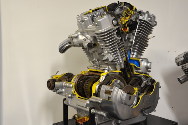 A sectioned motorcycle engine is displayed with detailed internal components visible. The machinery includes gears, pistons, and other mechanical parts, highlighted with bright yellow and blue accents to emphasize certain areas. The engine is mounted on a stand against a plain background.
