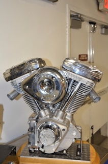 A chrome-plated motorcycle engine is displayed prominently. The engine features polished metal surfaces, prominent cylinder heads, and intricate mechanical components, resting on a wooden base. The surrounding environment is a plain indoor setting with part of a door and an exit sign visible in the background.
