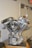 A chrome-plated motorcycle engine is displayed prominently. The engine features polished metal surfaces, prominent cylinder heads, and intricate mechanical components, resting on a wooden base. The surrounding environment is a plain indoor setting with part of a door and an exit sign visible in the background.