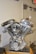 A chrome-plated motorcycle engine is displayed prominently. The engine features polished metal surfaces, prominent cylinder heads, and intricate mechanical components, resting on a wooden base. The surrounding environment is a plain indoor setting with part of a door and an exit sign visible in the background.