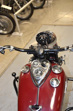 A vintage motorcycle dashboard with a round speedometer marked up to 120, set within a red tank adorned with chrome details. The handlebars are black with handgrips and controls visible. In the background, there are other motorcycle wheels and parts displayed.